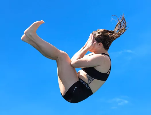 A person is displaying a classic Manu (V bomb) pose, against a blue sky background. They are mid-flight, yet to hit the water.