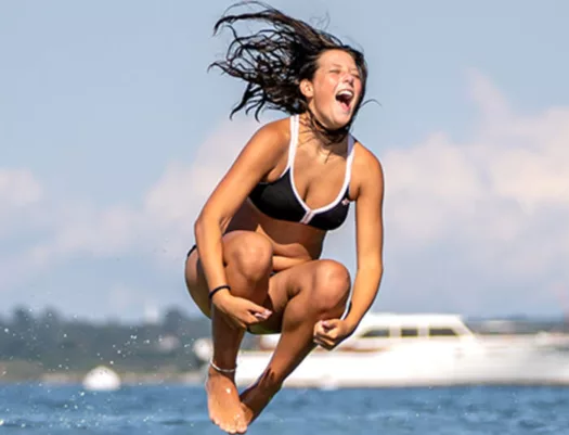 A person is smiling as they are about to enter the water in a Manu (bomb) pose and make a big splash.